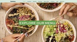 Top-down view of three colorful salad bowls with a bold green banner across the center reading EXPLORE OUR MENU, inviting users to view the menu.