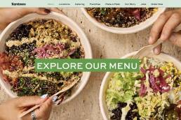 Top-down view of three colorful salad bowls with a bold green banner across the center reading EXPLORE OUR MENU, inviting users to view the menu.
